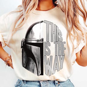 May include: Cream-colored sweatshirt featuring a black and white graphic of a Mandalorian helmet and the text "THIS IS THE WAY". The design is centered on the front of the shirt. The sweatshirt has long sleeves.