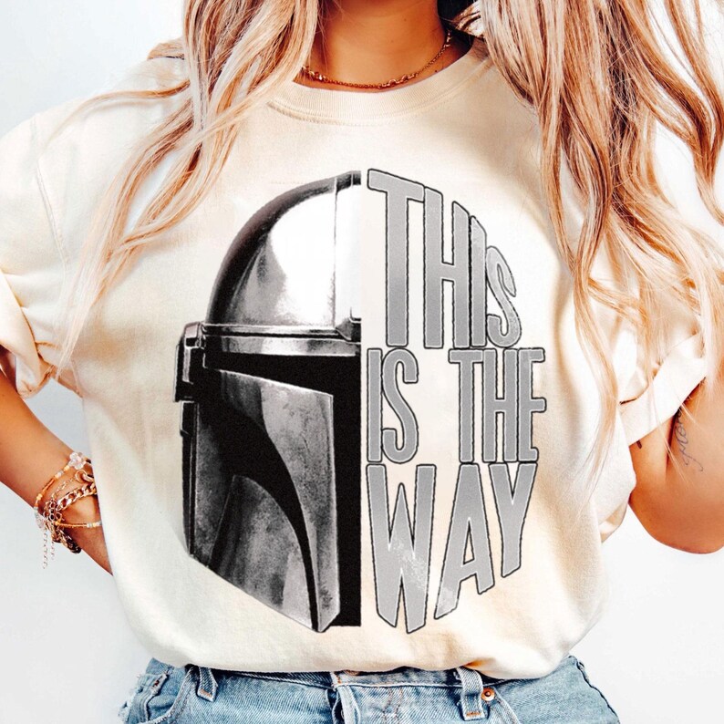 May include: Cream-colored sweatshirt featuring a black and white graphic of a Mandalorian helmet and the text "THIS IS THE WAY". The design is centered on the front of the shirt. The sweatshirt has long sleeves.