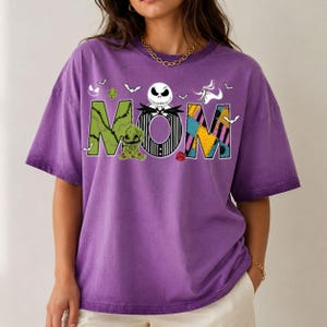 Puede incluir: Una camiseta morada de manga corta con la palabra MOM en letras grandes. La M es verde con puntadas, la O presenta una cara de Jack Skellington y la M es un patr&oacute;n de retazos. Un peque&ntilde;o fantasma y murci&eacute;lagos rodean el texto impreso.