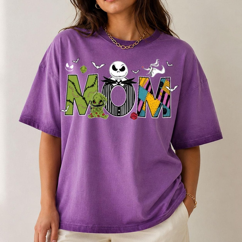Puede incluir: Una camiseta morada de manga corta con la palabra MOM en letras grandes. La M es verde con puntadas, la O presenta una cara de Jack Skellington y la M es un patr&oacute;n de retazos. Un peque&ntilde;o fantasma y murci&eacute;lagos rodean el texto impreso.
