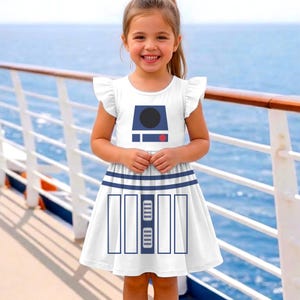 May include: White dress with a blue and black graphic design. The dress features a design on the bodice and skirt, with flutter sleeves. The dress is worn by a young person, with a background of water and a white railing.