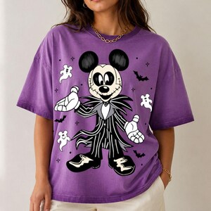 May include: A purple short-sleeve t-shirt featuring a graphic print of a character resembling Mickey Mouse dressed in a black and white pinstriped suit with a skull-like face, surrounded by small white ghost figures and black bat silhouettes.