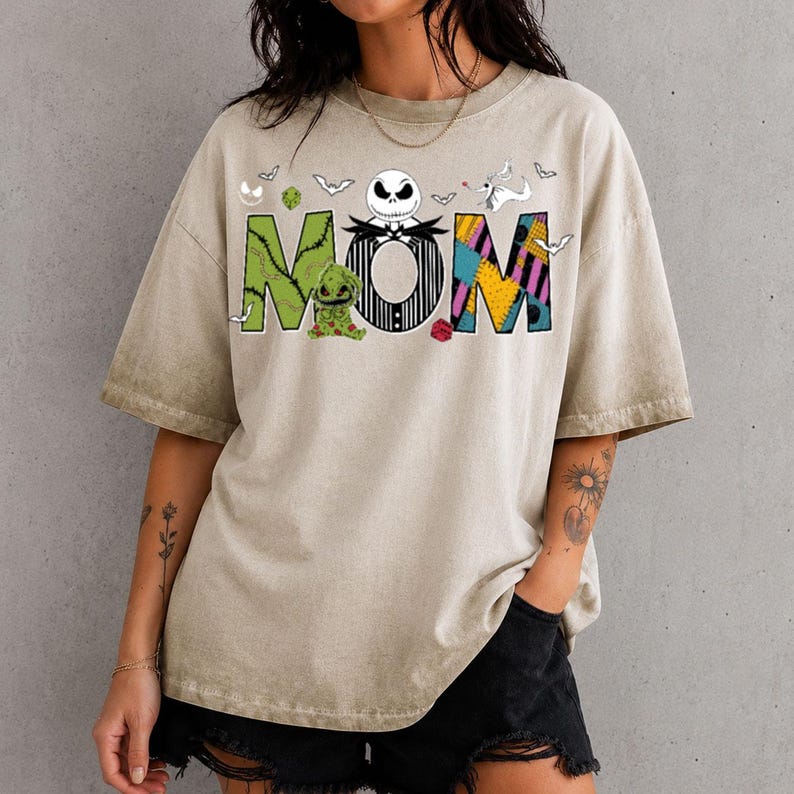 Puede incluir: Una camiseta beige de manga corta con la palabra MOM en letras grandes y estilizadas. La M es verde con costuras, la O presenta una cara de calavera blanca y la M es un mosaico de amarillo, morado y negro. Incluye peque&ntilde;os murci&eacute;lagos y un perro.