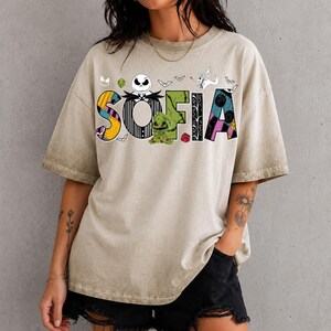 May include: A beige mineral-washed t-shirt featuring the name SOFIA in large, colorful letters. The design incorporates characters and motifs from The Nightmare Before Christmas, including Jack Skellington, Oogie Boogie, and Zero.