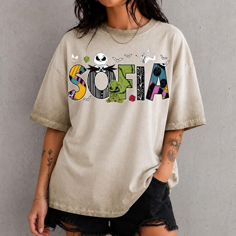 May include: A beige mineral-washed t-shirt featuring the name SOFIA in large, colorful letters. The design incorporates characters and motifs from The Nightmare Before Christmas, including Jack Skellington, Oogie Boogie, and Zero.
