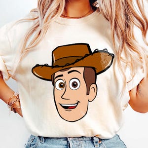 May include: Cream-colored t-shirt featuring a cartoon illustration of a smiling cowboy character with brown hair, a brown cowboy hat, and a red mouth. The character's face is outlined in black.