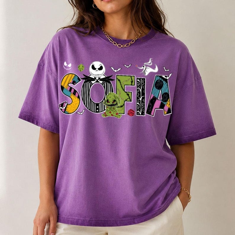 May include: A purple short-sleeve t-shirt featuring the name SOFIA in large, patterned letters. The design includes Jack Skellington, Oogie Boogie, and Zero from The Nightmare Before Christmas, along with small bats and a red die graphic.