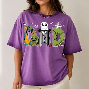 May include: A purple short-sleeve t-shirt featuring a graphic print of the word DAD. The letter D is a patchwork pattern, the letter A features a black and white striped suit with a skull character, and the letter D is green with stitches.