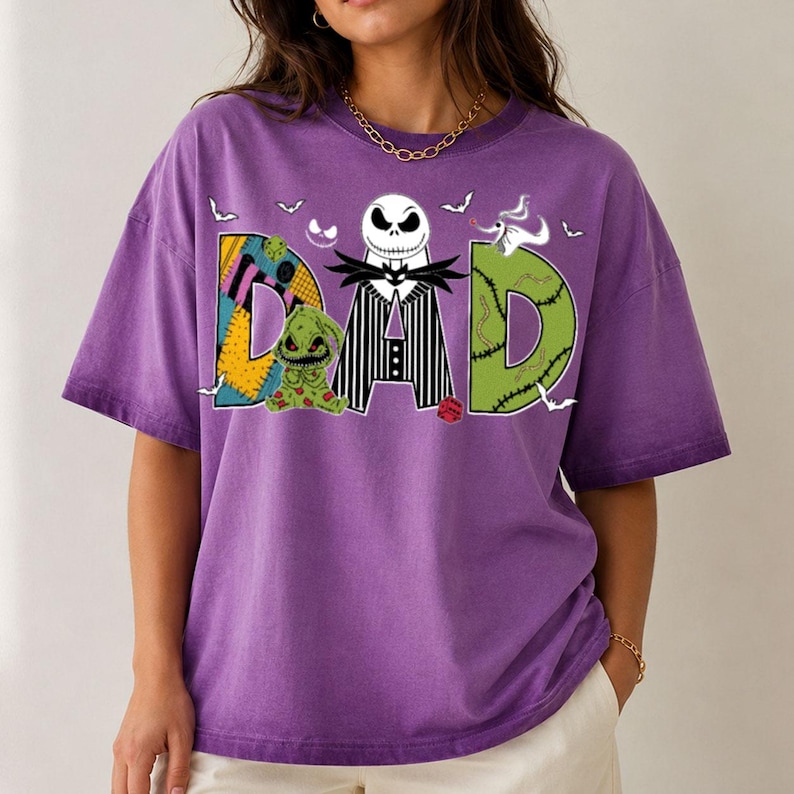 May include: A purple short-sleeve t-shirt featuring a graphic print of the word DAD. The letter D is a patchwork pattern, the letter A features a black and white striped suit with a skull character, and the letter D is green with stitches.