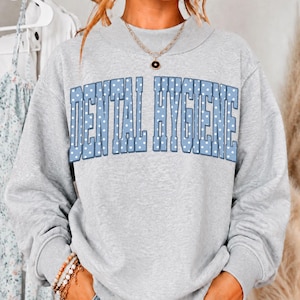 May include: A heather gray crewneck sweatshirt featuring the text DENTAL HYGIENE in large, light blue block letters with a white polka dot pattern. The garment has long sleeves, a ribbed neckline, and is worn with a gold chain necklace.