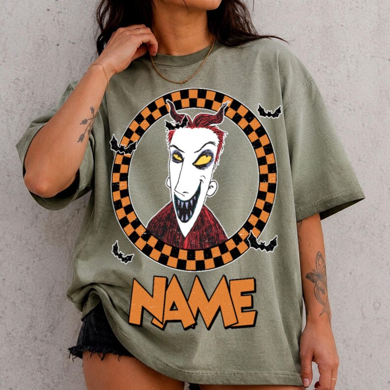 May include: An olive green t-shirt featuring a graphic of a character with red hair, yellow eyes, and a wide grin inside a black and orange checkered circle. Small black bats surround the circle, and the word NAME is printed in orange below.