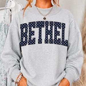 May include: A light gray crewneck sweatshirt featuring the word BETHEL in large, dark navy blue block letters with a white polka dot pattern. The garment has long sleeves with ribbed cuffs and a ribbed neckline, paired with a gold chain necklace.