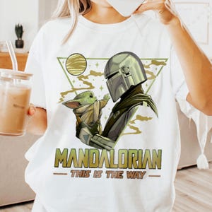 May include: White t-shirt featuring a graphic of The Mandalorian and Grogu. The design includes a helmeted figure and a small green creature, with the text "MANDALORIAN - THIS IS THE WAY" in green and gold.