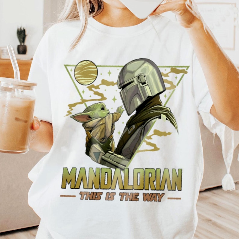 May include: White t-shirt featuring a graphic of The Mandalorian and Grogu. The design includes a helmeted figure and a small green creature, with the text "MANDALORIAN - THIS IS THE WAY" in green and gold.