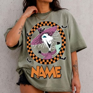 Puede incluir: Una camiseta verde oliva con un gr&aacute;fico de un personaje con sombrero morado puntiagudo y una m&aacute;scara dentro de un c&iacute;rculo a cuadros negro y naranja. Peque&ntilde;os murci&eacute;lagos negros rodean el c&iacute;rculo y la palabra NAME aparece en letras naranjas.