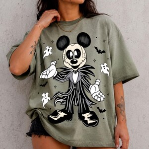 May include: An olive green short-sleeve t-shirt featuring a graphic print of a character resembling Mickey Mouse dressed in a black and white pinstriped suit, surrounded by small white ghost illustrations, black bats, and tiny star shapes.