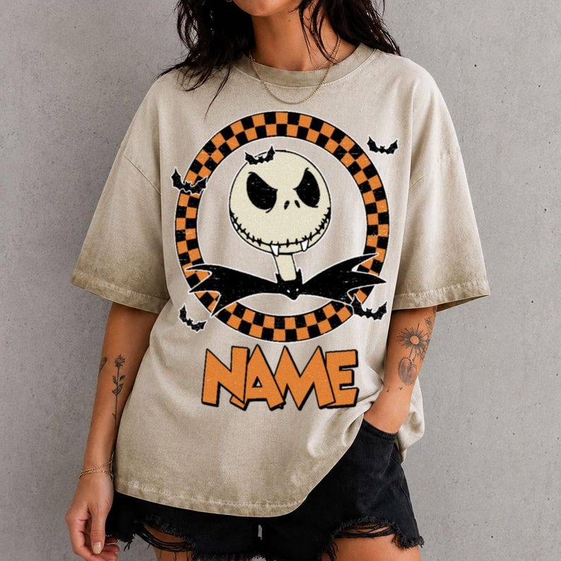 May include: A beige short-sleeve t-shirt featuring a graphic of a white skull character with a black bat bowtie inside a black and orange checkered circle. Small black bats surround the circle, and the word NAME is printed in orange below.