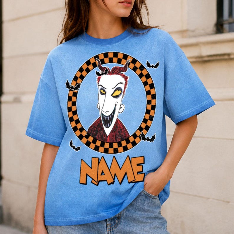 May include: A light blue short-sleeve t-shirt featuring a graphic of a character with red hair and sharp teeth inside a black and orange checkered circle. Small black bats surround the circle, and the word NAME is printed in orange below.