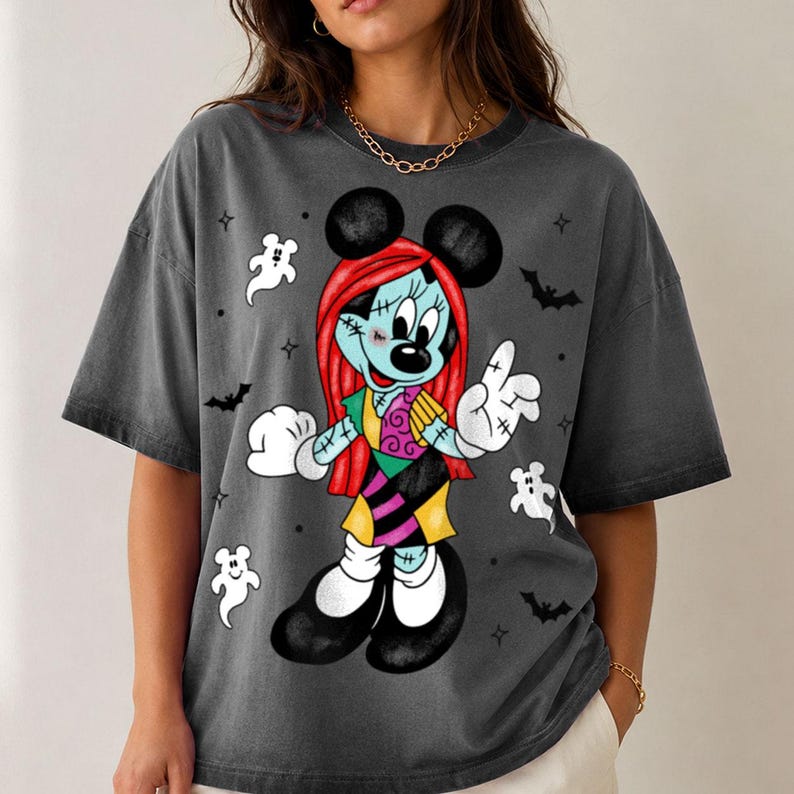 May include: A charcoal gray short-sleeve t-shirt featuring a graphic of a character resembling Minnie Mouse with red hair, stitched skin, and a patchwork dress. The design includes small white ghosts, black bats, and stars surrounding the figure.