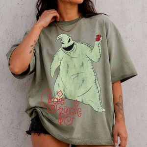 May include: An olive green short-sleeve t-shirt featuring a large graphic of a light green, stitched sack-like character holding red dice. Below the character, the red text reads Oogie Boogie in a stylized, cursive font with diamond accents.