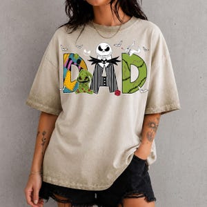 May include: A beige short-sleeve t-shirt featuring a graphic print that spells DAD. The letter D is a yellow and blue patchwork pattern, the A is a black and white striped suit with a skull head, and the D is green with stitched details.