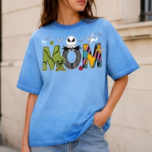 Puede incluir: Una camiseta de manga corta azul claro con la palabra MOM en letras grandes. La M es verde con costuras, la O es una cara de Jack Skellington a rayas blancas y negras, y la M es un patr&oacute;n de patchwork colorido con murci&eacute;lagos y Zero.