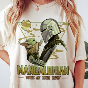 May include: Cream-colored t-shirt featuring a graphic of The Mandalorian and Grogu. The Mandalorian is depicted in his helmet, facing Grogu. The text "MANDALORIAN - THIS IS THE WAY" is printed in green and gold.