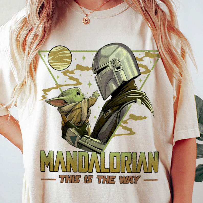 May include: Cream-colored t-shirt featuring a graphic of The Mandalorian and Grogu. The Mandalorian is depicted in his helmet, facing Grogu. The text "MANDALORIAN - THIS IS THE WAY" is printed in green and gold.