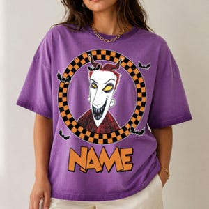 May include: A purple short-sleeve t-shirt featuring a graphic of a character with red hair and sharp teeth inside a black and orange checkered circle. Small black bats surround the circle, and the word NAME is printed in orange below the graphic.