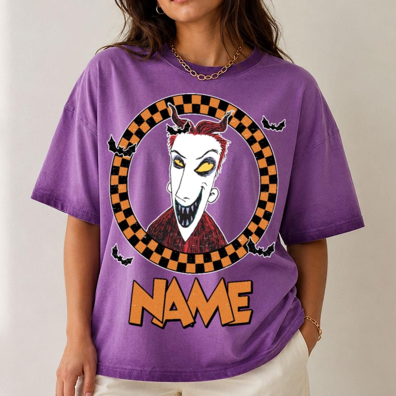 May include: A purple short-sleeve t-shirt featuring a graphic of a character with red hair and sharp teeth inside a black and orange checkered circle. Small black bats surround the circle, and the word NAME is printed in orange below the graphic.