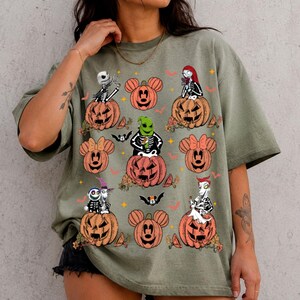 May include: An olive green short-sleeve t-shirt featuring a graphic print of various characters from The Nightmare Before Christmas sitting on orange jack-o'-lanterns shaped like Mickey Mouse ears, interspersed with small bats and stars.