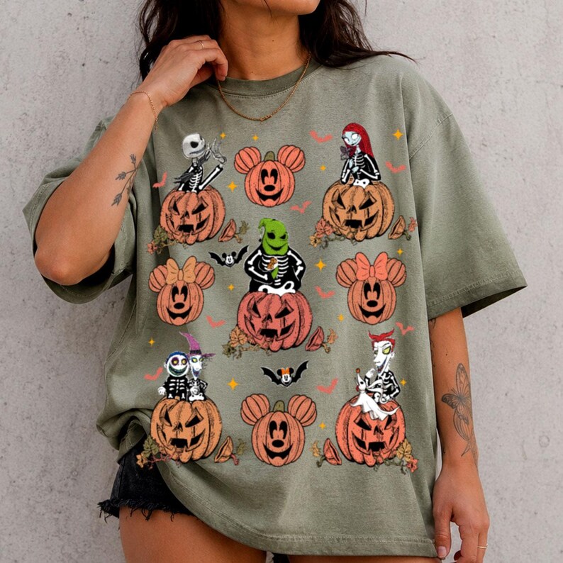 May include: An olive green short-sleeve t-shirt featuring a graphic print of various characters from The Nightmare Before Christmas sitting on orange jack-o'-lanterns shaped like Mickey Mouse ears, interspersed with small bats and stars.