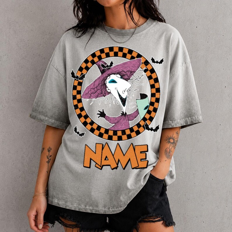 Puede incluir: Una camiseta gris con efecto lavado al &aacute;cido que presenta un gr&aacute;fico de un personaje con sombrero morado dentro de un c&iacute;rculo a cuadros naranja y negro. Peque&ntilde;os murci&eacute;lagos negros rodean el c&iacute;rculo. Debajo, la palabra NAME en letras naranjas.