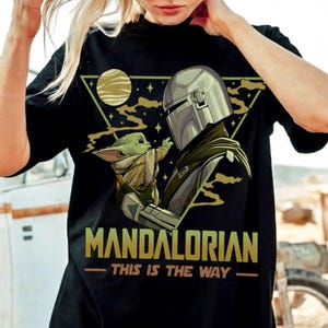 May include: Black t-shirt featuring a graphic of The Mandalorian and Grogu, with the text "MANDALORIAN - THIS IS THE WAY" in gold. The design includes a helmeted figure and a small green creature against a backdrop of a planet and stars.