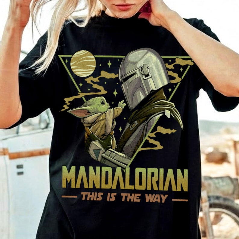 May include: Black t-shirt featuring a graphic of The Mandalorian and Grogu, with the text "MANDALORIAN - THIS IS THE WAY" in gold. The design includes a helmeted figure and a small green creature against a backdrop of a planet and stars.
