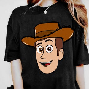 May include: Black t-shirt featuring a cartoon illustration of a smiling cowboy character with a brown hat and hair, and light skin. The character's eyes are brown, and the shirt is a solid black color.