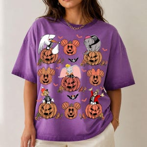 May include: A purple short-sleeve t-shirt featuring a graphic print of various Disney characters, including Dumbo and Mickey Mouse, sitting on top of orange jack-o'-lantern pumpkins. The design also includes small orange bats and autumn leaves.