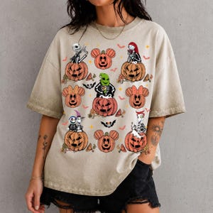 May include: A beige short-sleeve t-shirt featuring a graphic print of various characters from The Nightmare Before Christmas sitting in or near orange jack-o'-lanterns, interspersed with small orange bats and stars on the front.