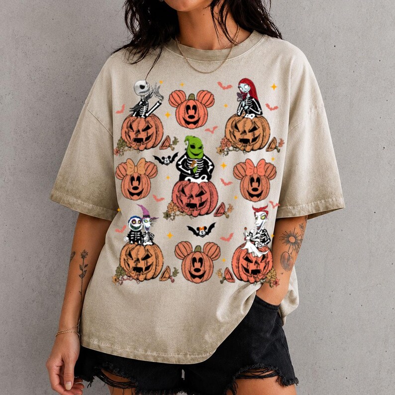 May include: A beige short-sleeve t-shirt featuring a graphic print of various characters from The Nightmare Before Christmas sitting in or near orange jack-o'-lanterns, interspersed with small orange bats and stars on the front.