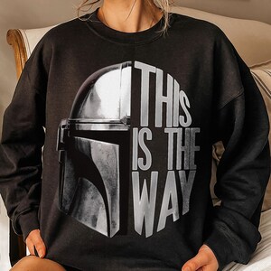 May include: Black sweatshirt featuring a graphic of a Mandalorian helmet and the text "THIS IS THE WAY" in white. The design is centered on the front of the long-sleeved top, creating a bold statement. The sweatshirt is a comfortable, casual wear item.