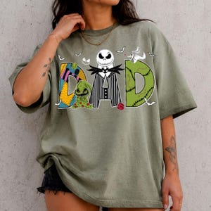 May include: An olive green t-shirt featuring the word DAD in large letters. The letter D is a patchwork pattern, the A is a black and white striped suit with a skull head, and the D is green with stitches. A small green creature is included.