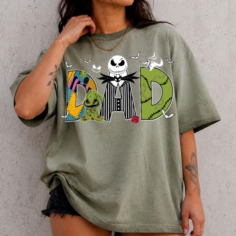 May include: An olive green t-shirt featuring the word DAD in large letters. The letter D is a patchwork pattern, the A is a black and white striped suit with a skull head, and the D is green with stitches. A small green creature is included.