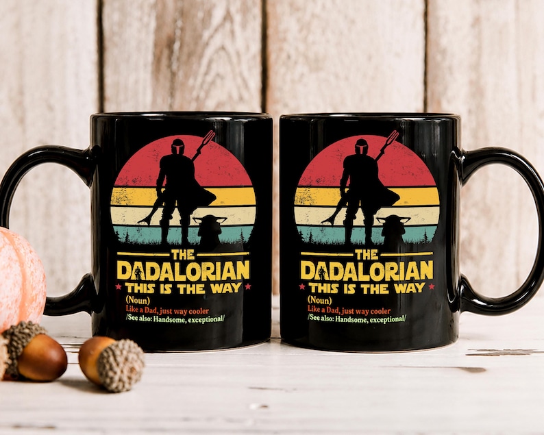 May include: Two black ceramic mugs with a graphic design featuring a silhouette of a figure with a staff, a baby-like creature, and the text "THE DADALORIAN THIS IS THE WAY". The design is set against a retro sunset.