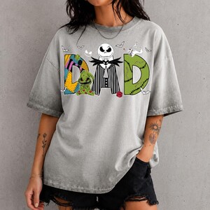 May include: A grey short-sleeve t-shirt featuring a graphic print that spells DAD. The letter D is a yellow and blue patchwork pattern, the letter A is a black and white striped suit with a skull head, and the letter D is green with stitches.