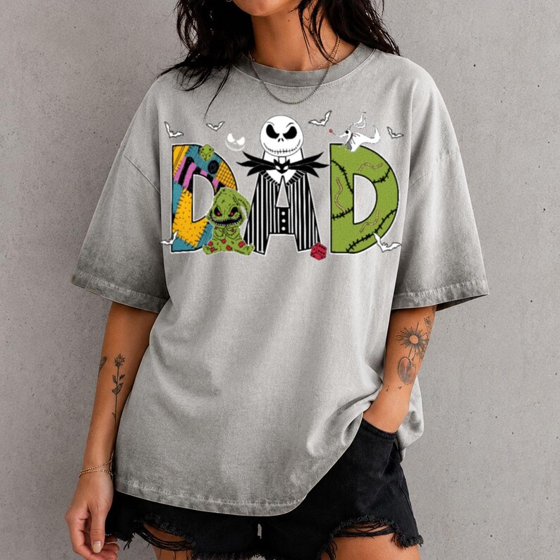May include: A grey short-sleeve t-shirt featuring a graphic print that spells DAD. The letter D is a yellow and blue patchwork pattern, the letter A is a black and white striped suit with a skull head, and the letter D is green with stitches.