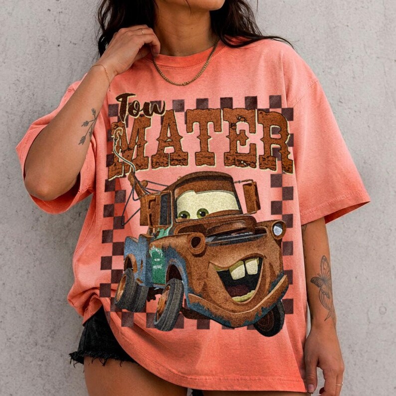 May include: A peach-colored short-sleeve t-shirt featuring a graphic print of a rusty brown tow truck character with large eyes and a wide smile. The text "Tow Mater" is printed in a distressed brown font above the truck over a brown checkerboard.