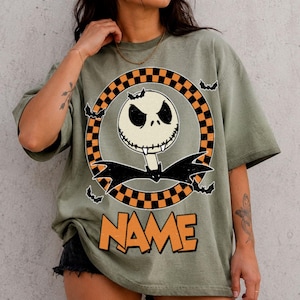 May include: An olive green t-shirt featuring a graphic of a white skull character with a stitched mouth and black bat wings. The character is centered within an orange and black checkered circle, surrounded by small black bats, with the word NAME below.