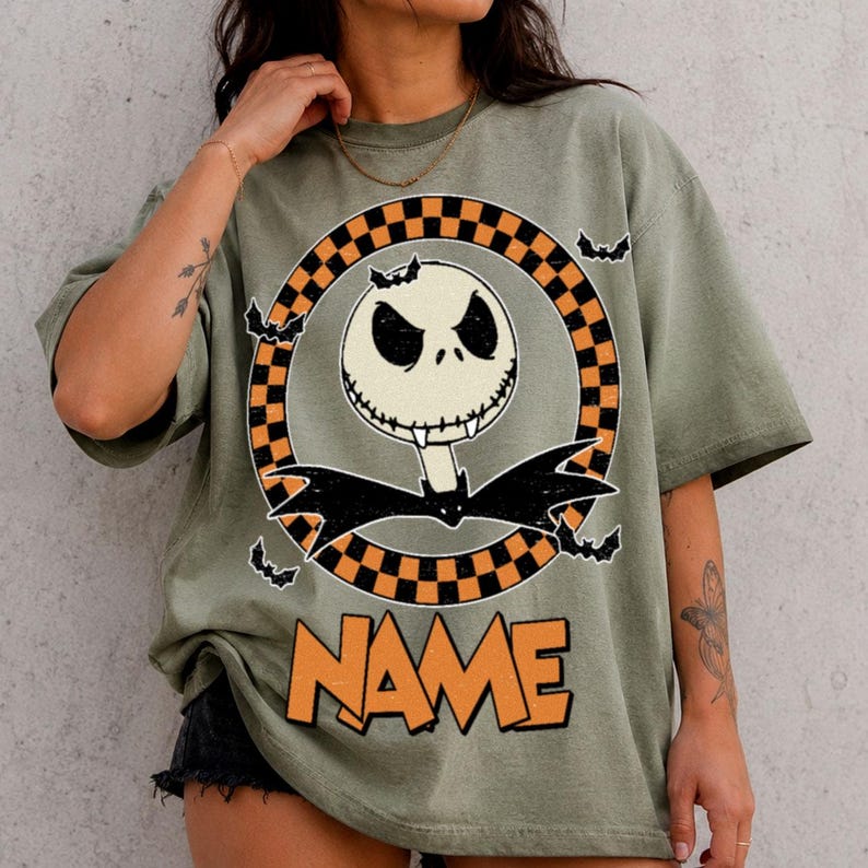 May include: An olive green t-shirt featuring a graphic of a white skull character with a stitched mouth and black bat wings. The character is centered within an orange and black checkered circle, surrounded by small black bats, with the word NAME below.