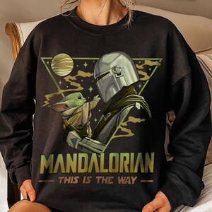 May include: Black sweatshirt featuring a graphic of The Mandalorian and Grogu. The design includes a helmeted figure and a small green creature, with the text "MANDALORIAN" and "THIS IS THE WAY" in gold lettering.