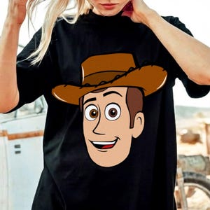 May include: Black t-shirt featuring a cartoon illustration of a cowboy with a brown hat and a smiling face. The cowboy's face is tan with brown hair and eyes. The shirt is a crew neck style.